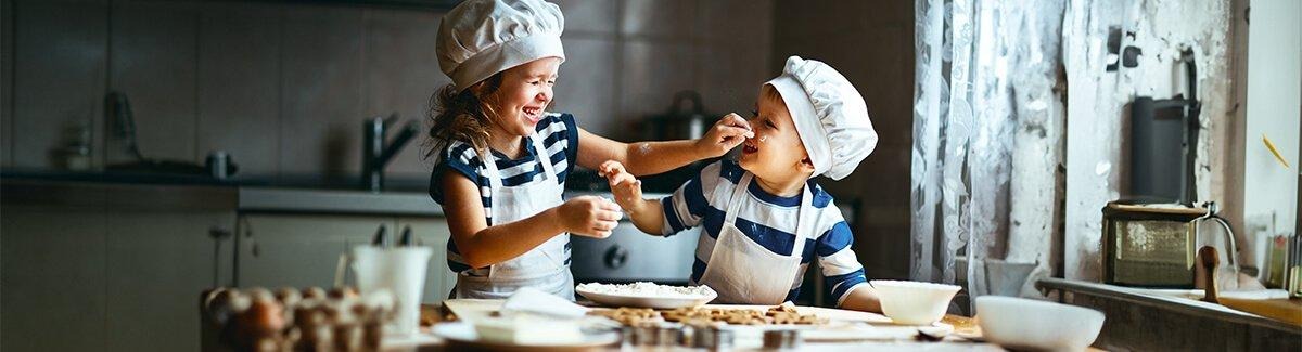 Dos niños con gorros y delantales de chef jugando con harina en la cocina, sonriendo y divirtiéndose.