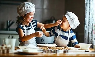 Dos niños con gorros y delantales de chef jugando con harina en la cocina, sonriendo y divirtiéndose.