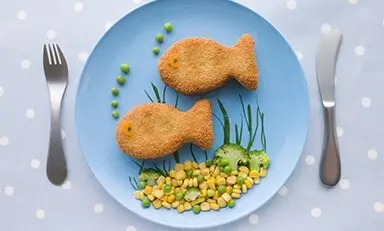 Tostada con queso y verduras formando la figura de un pez, en un plato blanco con fondo azul. El texto en español promueve el desarrollo intelectual de los niños con omega 6 y 3. Se muestra una lata de PediaSure en la esquina inferior derecha.