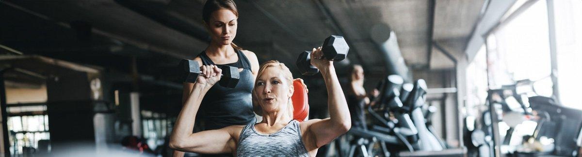 Mujer adulta en ropa deportiva, realizado levantamiento de pesas con la supervisión de alguien en el gimnasio