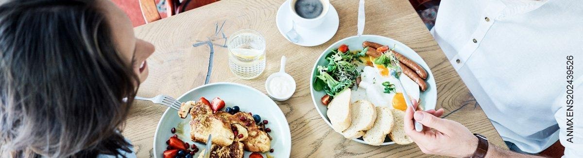 Pareja de adultos comiendo un desayuno saludable de: huevos, salchicha, pan, fresas, arándanos, tostadas francesas y más.