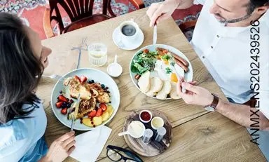 Pareja de adultos comiendo un desayuno saludable de: huevos, salchicha, pan, fresas, arándanos, tostadas francesas y más.