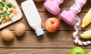 mesa de elementos de un estilo de vida saludable, como: mancuerdas, metro, una bebida y alimentos saludables como: frutas, verduras, huevos.