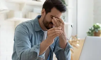 Hombre sosteniendo sus sienes con expresión de dolor, sujetando sus gafas con la otra mano