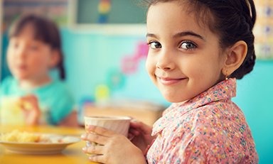 Niño sosteniendo una taza, con el rostro borroso por privacidad. El niño lleva una camisa con patrón floral y está sentado en una mesa con un plato de comida al fondo
