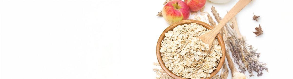 Cuenco de madera lleno de avena enrollada. Dentro del cuenco hay una cuchara de madera. Alrededor del cuenco hay avena esparcida, espigas de trigo y dos manzanas.