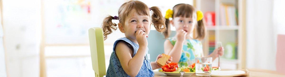 Dos niños sentados a la mesa, comiendo una comida. Los rostros de los niños están difuminados por privacidad. La mesa tiene platos con alimentos como verduras en rodajas y un sándwich, y también hay un vaso de agua.