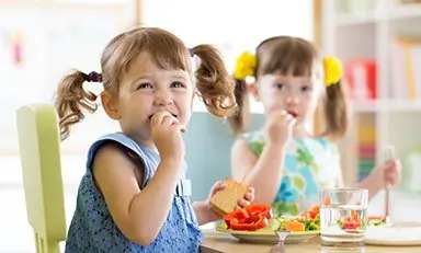 Dos niños sentados a la mesa, comiendo una comida. Los rostros de los niños están difuminados por privacidad. La mesa tiene platos con alimentos como verduras en rodajas y un sándwich, y también hay un vaso de agua.