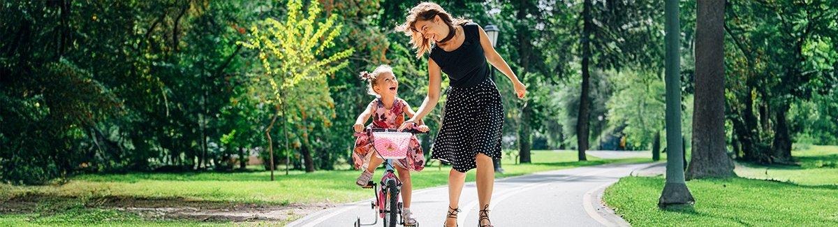 Niño montando una bicicleta con la ayuda de un adulto en un entorno al aire libre, con vegetación de fondo. El adulto viste una blusa negra y una falda con lunares, mientras que el niño lleva un vestido floral.