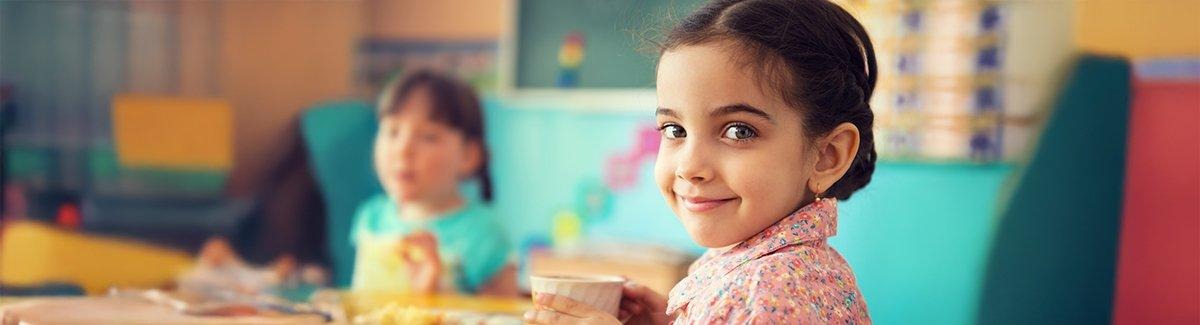 Niño sosteniendo una taza, con el rostro borroso por privacidad. El niño lleva una camisa con patrón floral y está sentado en una mesa con un plato de comida al fondo