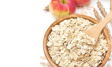 Cuenco de madera lleno de avena enrollada. Dentro del cuenco hay una cuchara de madera. Alrededor del cuenco hay avena esparcida, espigas de trigo y dos manzanas.