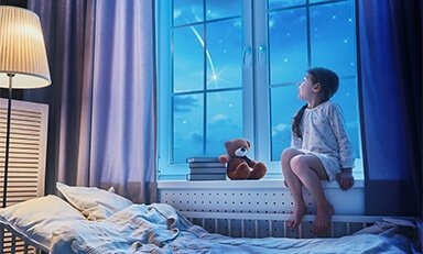 Niño sentado en un alféizar, mirando el cielo nocturno con una estrella fugaz visible. El niño lleva pijama y está acompañado por un oso de peluche y una pila de libros.