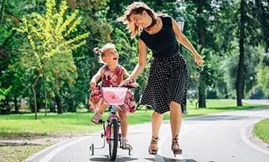 Niño montando una bicicleta con la ayuda de un adulto en un entorno al aire libre, con vegetación de fondo. El adulto viste una blusa negra y una falda con lunares, mientras que el niño lleva un vestido floral.