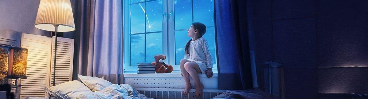 Niño sentado en un alféizar, mirando el cielo nocturno con una estrella fugaz visible. El niño lleva pijama y está acompañado por un oso de peluche y una pila de libros.