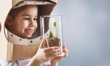 Niño sosteniendo una pequeña planta en un contenedor de vidrio. El niño lleva una caja de cartón con agujeros recortados para la cara y el cuello, semejante a un casco espacial.