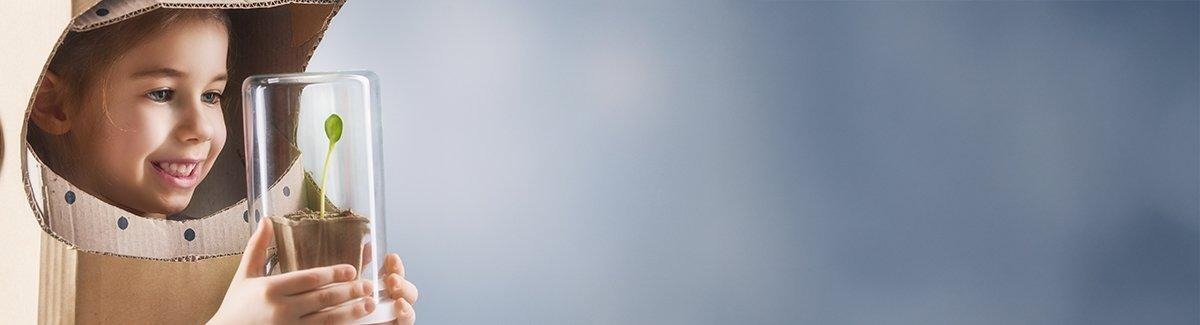 Niño sosteniendo una pequeña planta en un contenedor de vidrio. El niño lleva una caja de cartón con agujeros recortados para la cara y el cuello, semejante a un casco espacial.