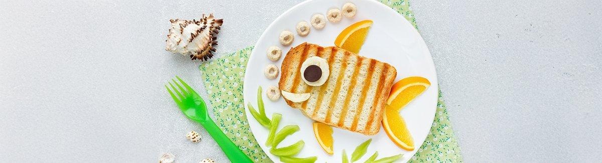 Rebanada de pan tostado en un plato blanco sobre un fondo azul. La tostada está cubierta con queso, rodajas de albaricoque, una rodaja de aceituna negra y una rodaja de aceituna verde.