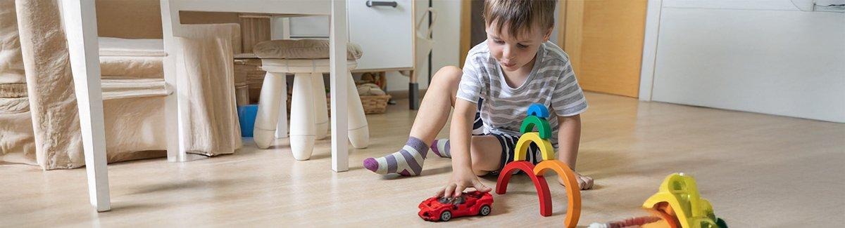 Un niño tumbado en el suelo, jugando con un coche de juguete. El niño lleva una camisa de cuadros y calcetines azules. El fondo muestra un entorno interior borroso con un sofá de color claro y una mesa