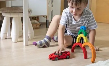 Un niño tumbado en el suelo, jugando con un coche de juguete. El niño lleva una camisa de cuadros y calcetines azules. El fondo muestra un entorno interior borroso con un sofá de color claro y una mesa