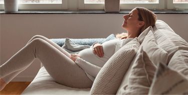 Mujer embarazada, sentada en un sofá con las piernas dobladas, acariciando su barriga con una sonrisa y los ojos cerrados”