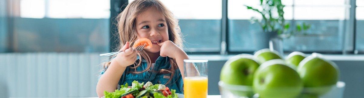 alt="Niña sonríe tímidamente mientras come un plato de verduras con jugo de naranja"