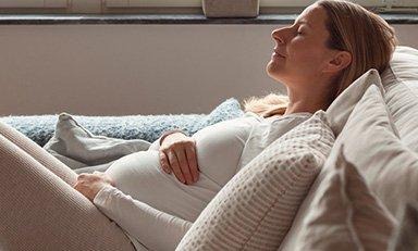 Mujer embarazada, sentada en un sofá con las piernas dobladas, acariciando su barriga con una sonrisa y los ojos cerrados
