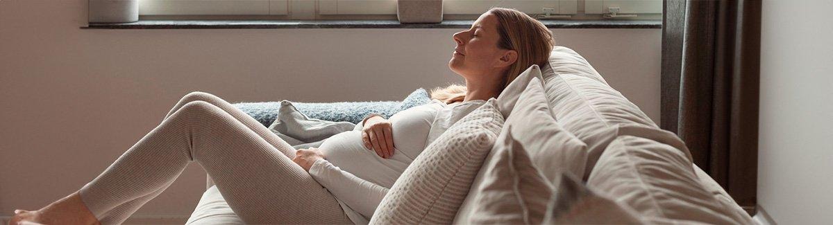 Mujer embarazada, sentada en un sofá con las piernas dobladas, acariciando su barriga con una sonrisa y los ojos cerrados