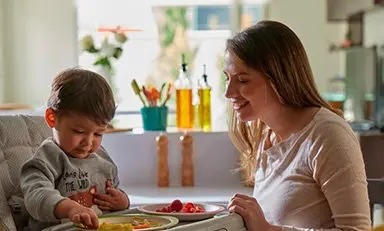 Niño sentado en una silla comedor cogiendo una porción de un plato con fruta junto con su mamá frente a él, sonriéndole