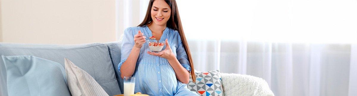Mujer embarazada en el sofá comiendo un bowl de fruta y avena, junto con su desayuno en una bandeja con Similac ® Mamá