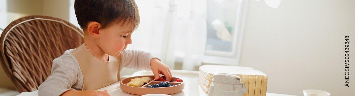 Niño de dos años sentado cogiendo una porción de un plato con banano, frambuesas y arándanos