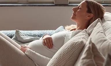 Mujer embarazada, sentada en un sofá con las piernas dobladas, acariciando su barriga con una sonrisa y los ojos cerrados”