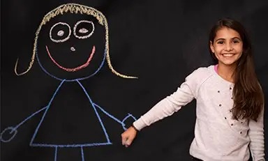 Niño de pie frente a una pizarra con dibujos de un alienígena y una nave espacial. El niño señala hacia los dibujos, imitando una acción de disparo.