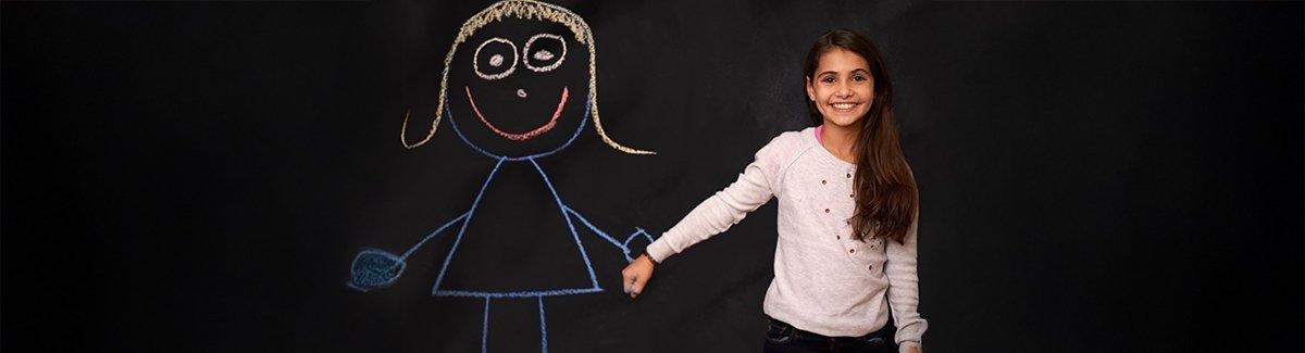 Niño de pie frente a una pizarra con dibujos de un alienígena y una nave espacial. El niño señala hacia los dibujos, imitando una acción de disparo.